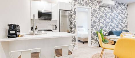 A modern kitchen with white cabinets, a stainless steel refrigerator, and a coffee machine. There is a dining area with a yellow chair and a table.
