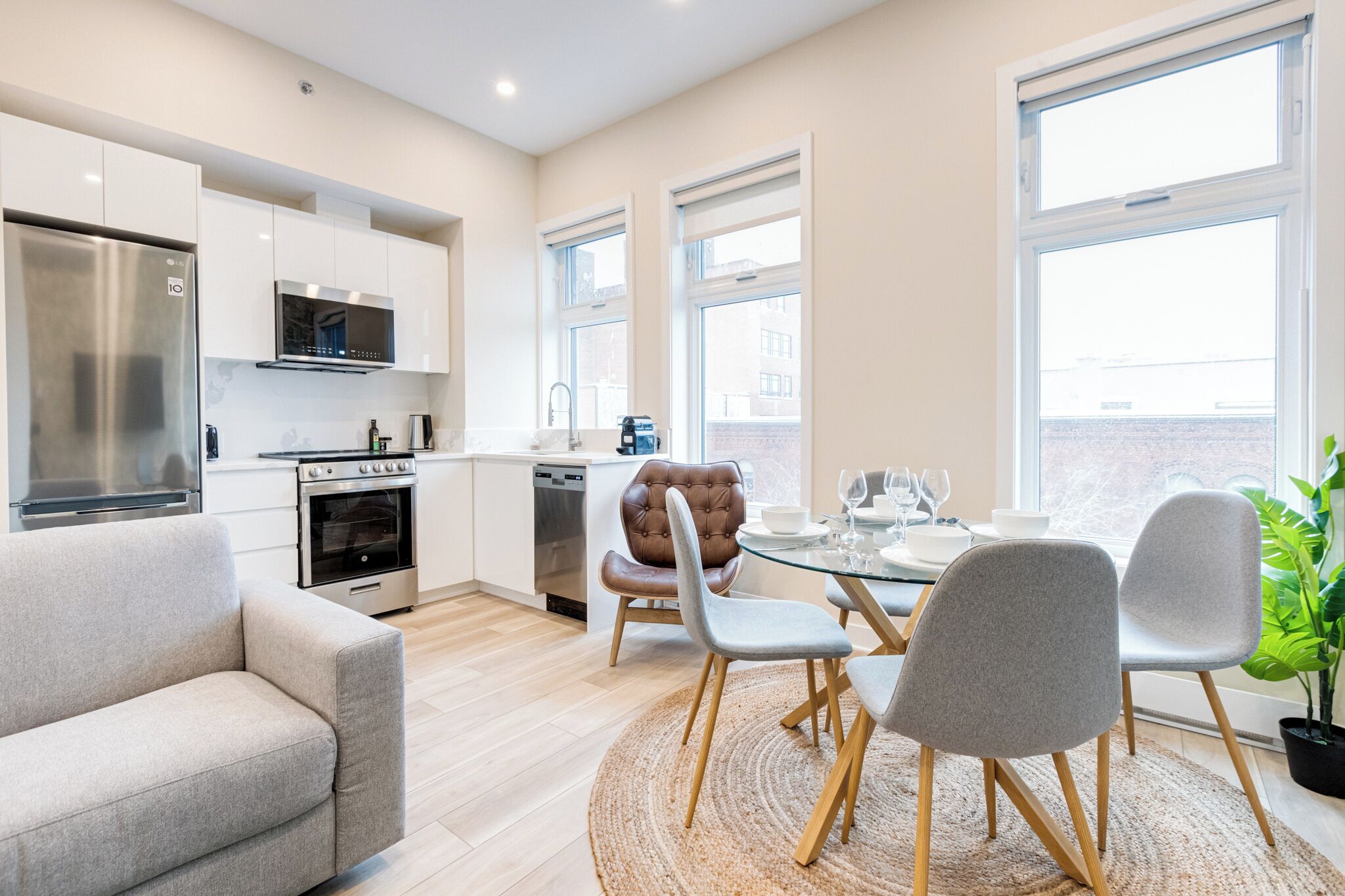 Una cocina moderna con electrodomésticos de acero inoxidable, una zona de comedor con mesa de vidrio y sillas grises, y ventanas grandes que ofrecen luz natural.