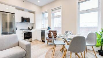 Una cocina moderna con electrodomésticos de acero inoxidable, una zona de comedor con mesa de vidrio y sillas grises, y ventanas grandes que ofrecen luz natural.