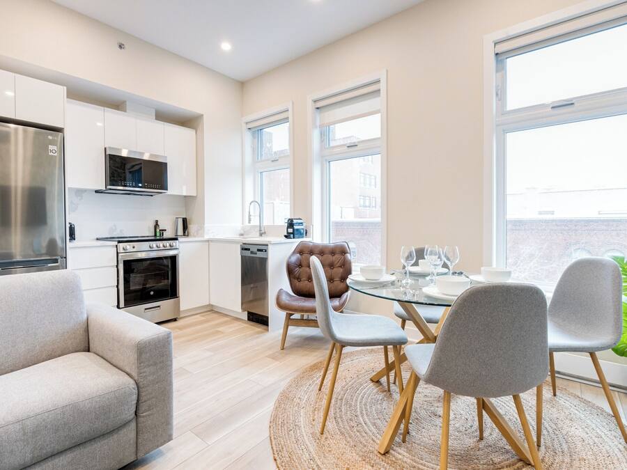 Una cocina moderna con electrodomésticos de acero inoxidable, una zona de comedor con mesa de vidrio y sillas grises, y ventanas grandes que ofrecen luz natural.