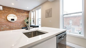 Una cucina moderna con un piano di lavoro bianco, lavastoviglie integrata, parete in mattoni e un'ampia finestra.