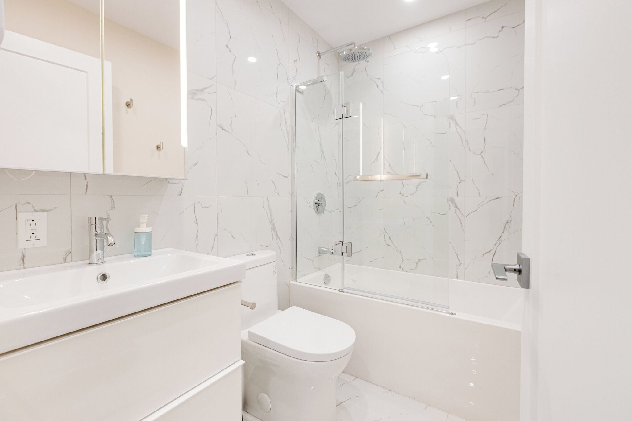 Une salle de bain moderne avec un pare-chocs en marbre blanc, une douche à l’italienne en verre, un lavabo blanc et des toilettes.