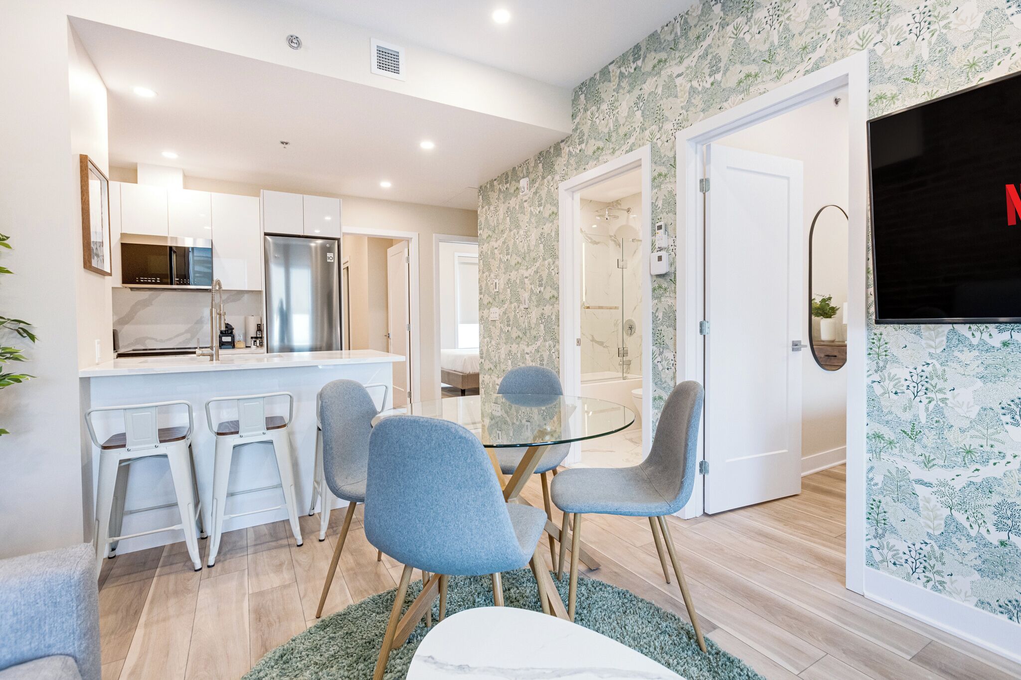 Une cuisine moderne avec des meubles blancs, un réfrigérateur en acier inoxydable et une table à manger en verre, assortie de chaises bleues.