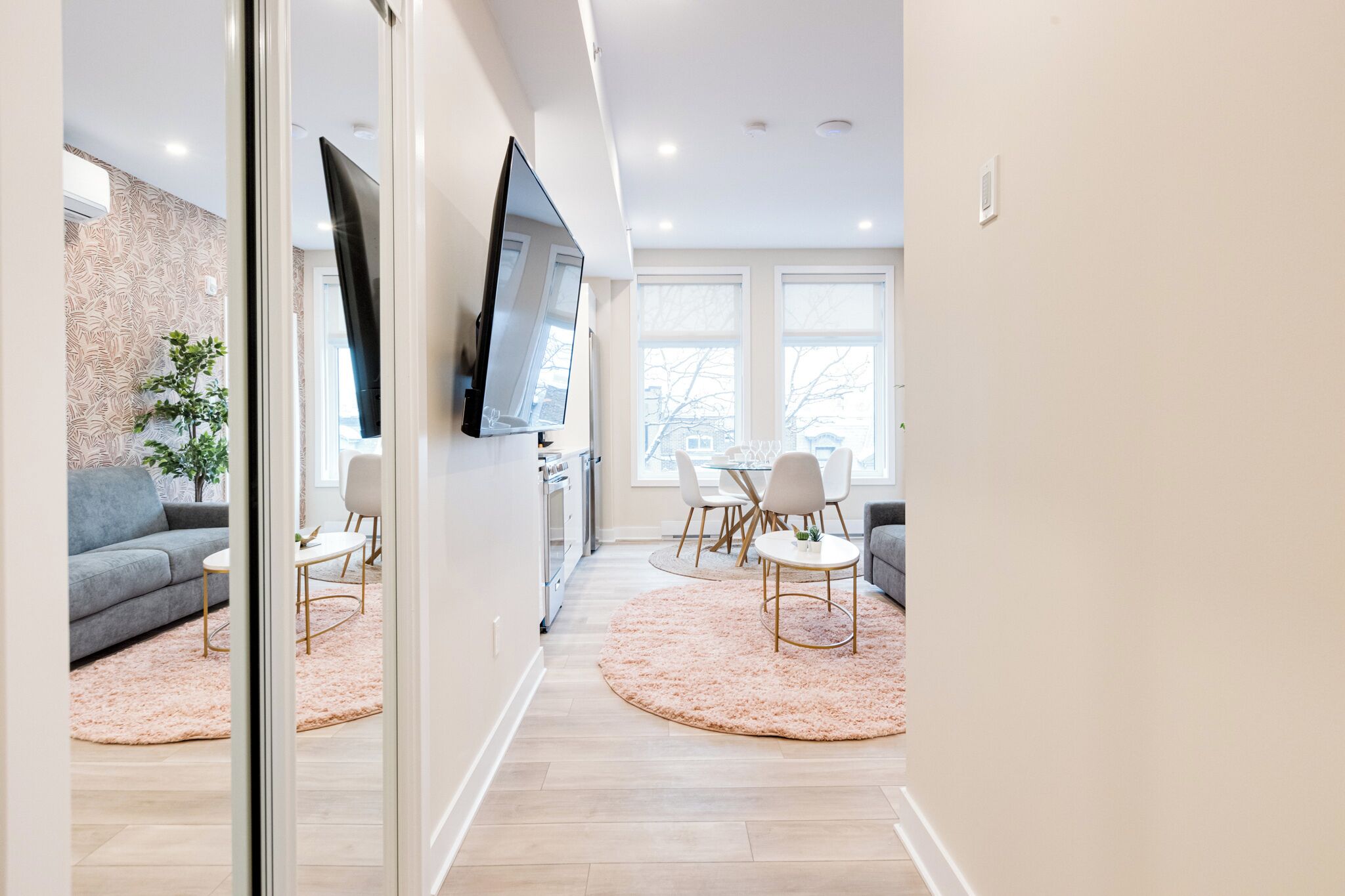 A modern living room with a grey sofa, a TV mounted on the wall, and a dining area with a table and chairs.