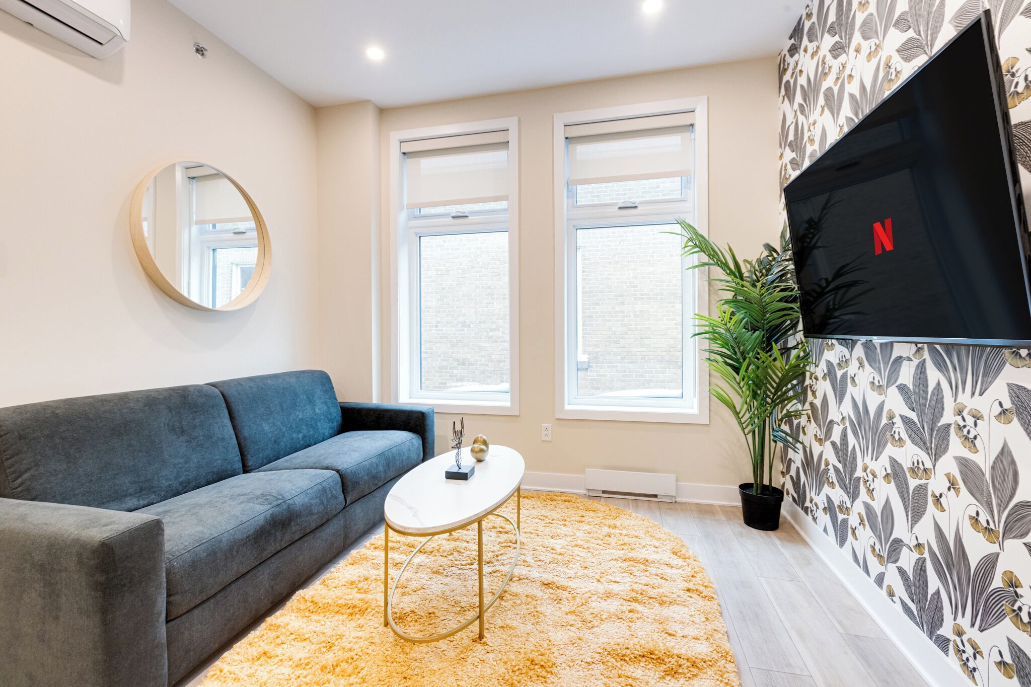 A living room with a grey sofa, a round mirror, a white coffee table, and a large TV on the wall.
