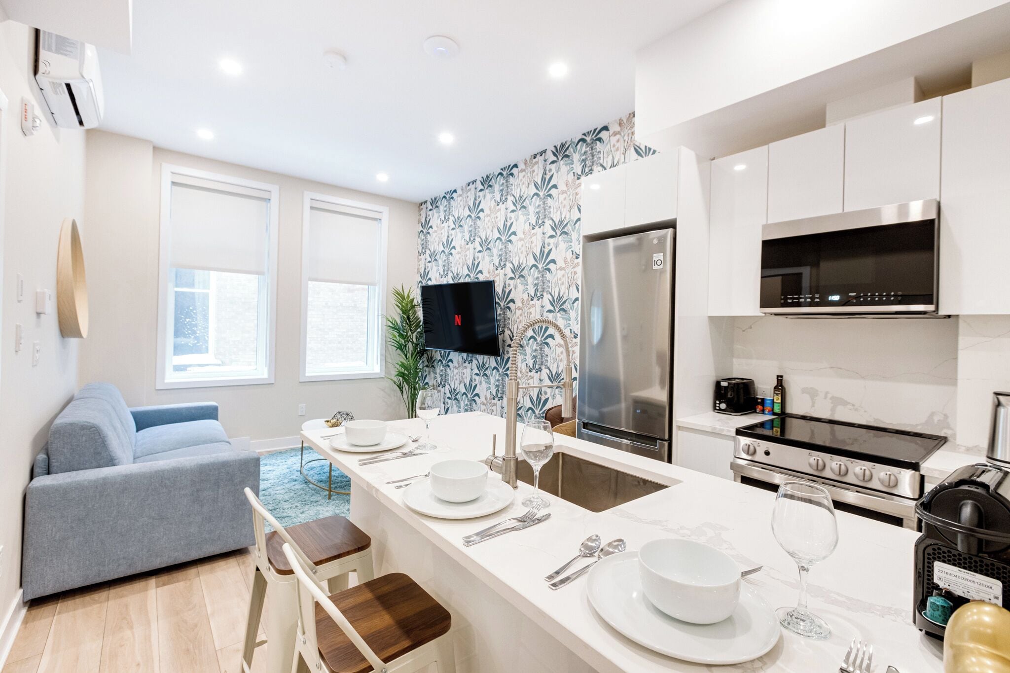 A modern kitchen with white cabinets, a stainless steel refrigerator, a built-in oven, and a microwave. There is a dining area with a table set for two, and a gray sofa in the living area.