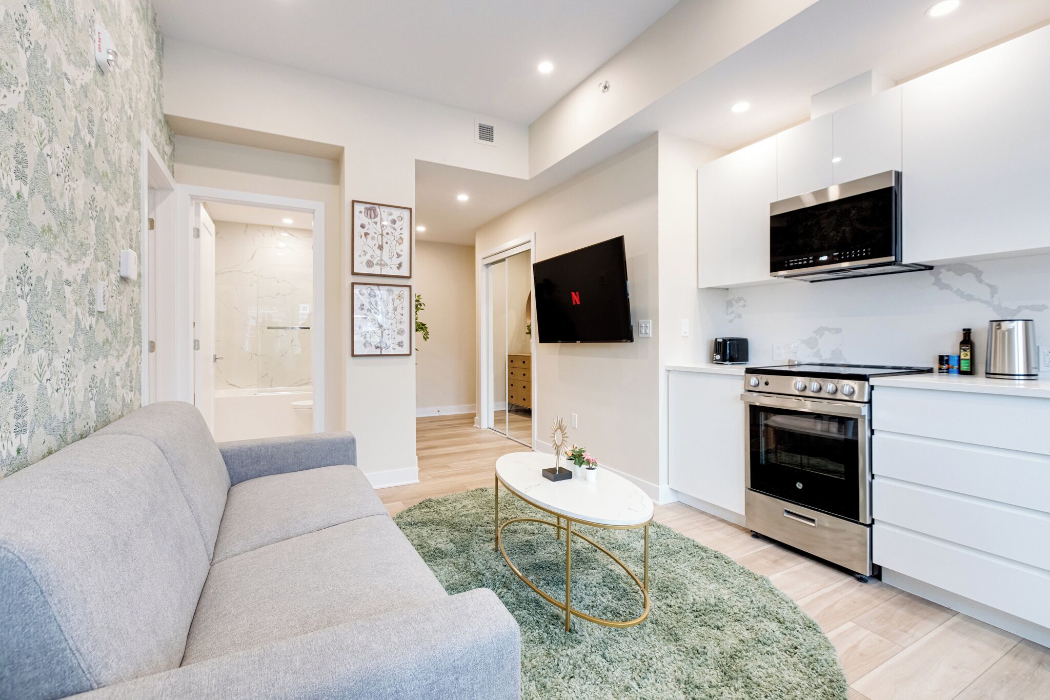 A modern living room with a grey sofa, a small white coffee table, and a green rug. It leads to a kitchen with white cabinets, a stainless steel oven, and a flat-screen TV mounted above the stove.
