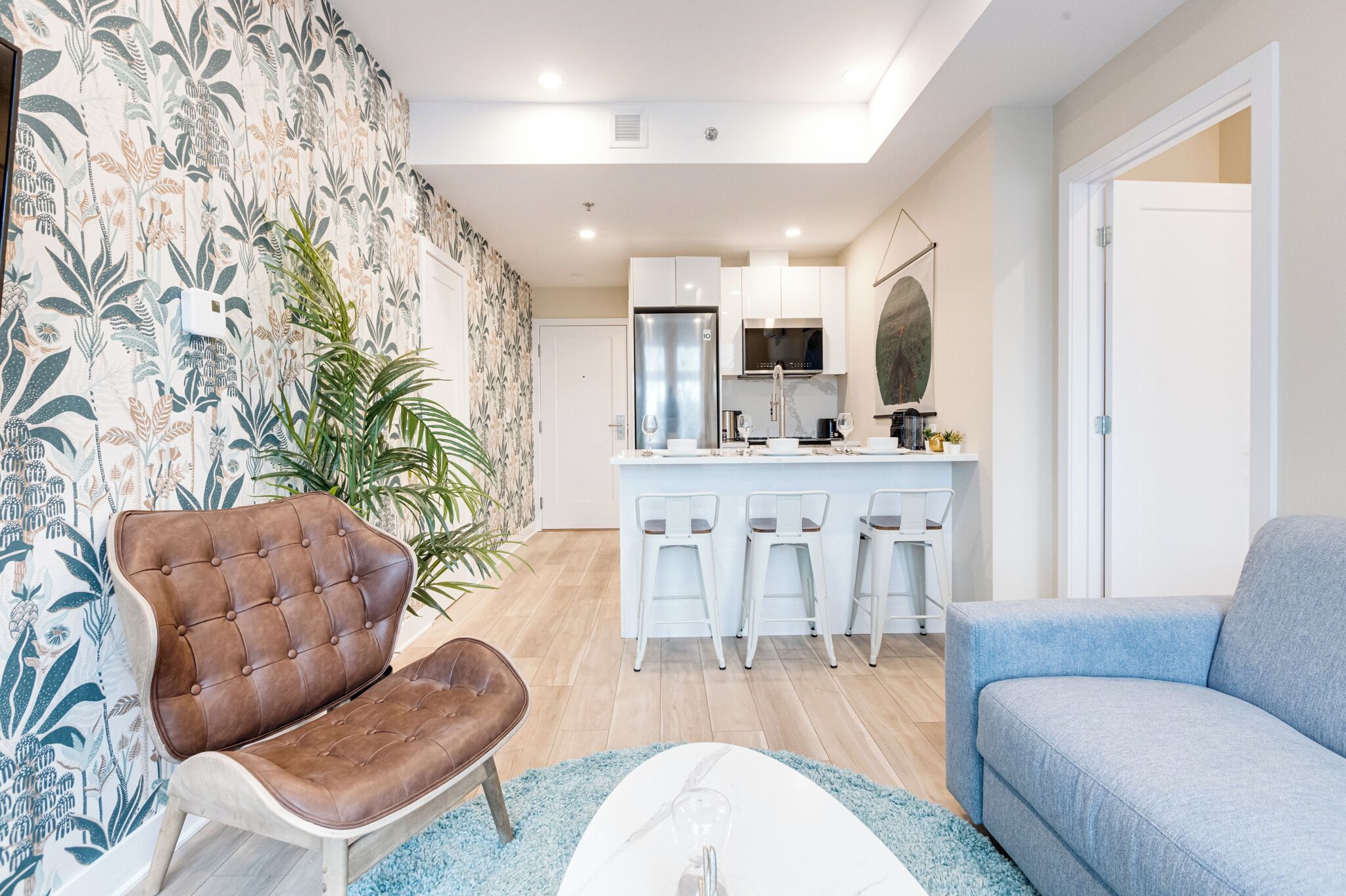 Un salon moderne avec un canapé, un fauteuil en cuir marron, un coin repas avec un bar et une cuisine en arrière-plan.