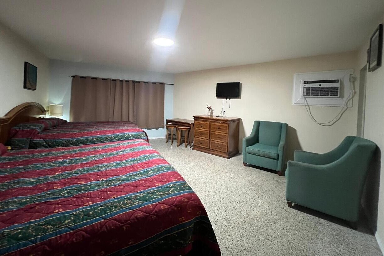 A hotel room with two beds, a desk, a chair, a dresser, a television, and an air conditioning unit.