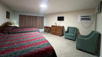 A hotel room with two beds, a desk, a chair, a dresser, a television, and an air conditioning unit.