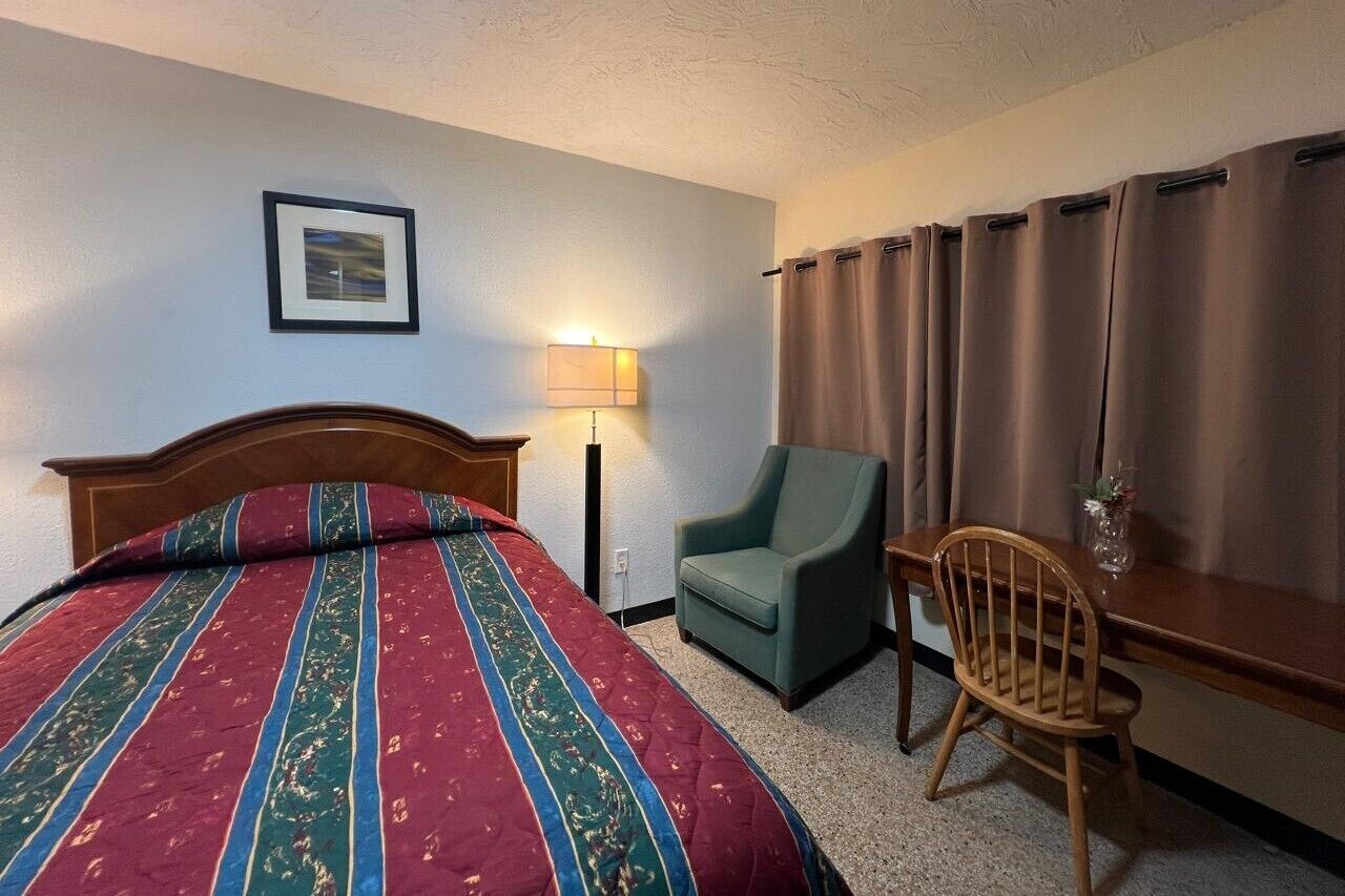 A hotel room with a bed, a desk, a chair, a lamp, and a picture on the wall.