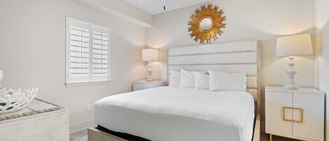 Un dormitorio con una cama, un ventilador de techo, una mesita de noche, una lámpara, un espejo decorativo y una pared blanca con una ventana.