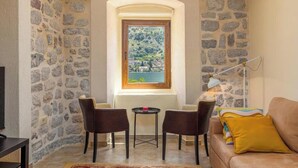 Une pièce chaleureuse aux murs de pierre, avec une fenêtre à cadre en bois, deux chaises, une petite table et un canapé agrémenté de coussins.