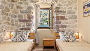 Une pièce aux murs de pierre, avec deux lits, une petite table et une fenêtre donnant sur une colline.