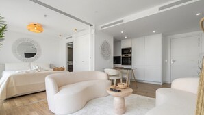 Quarto moderno com cama grande, sofá, mesa pequena e cozinha ao fundo.