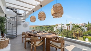Terraço no telhado com mesa de madeira posta para seis pessoas, rodeada por cadeiras de vime, sob três luminárias pendentes trançadas. Uma escada em espiral é visível ao fundo.