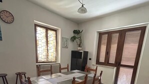 Un comedor con una mesa rectangular, seis sillas de madera, dos lámparas colgantes, una planta en maceta, un radiador y una ventana con persianas.