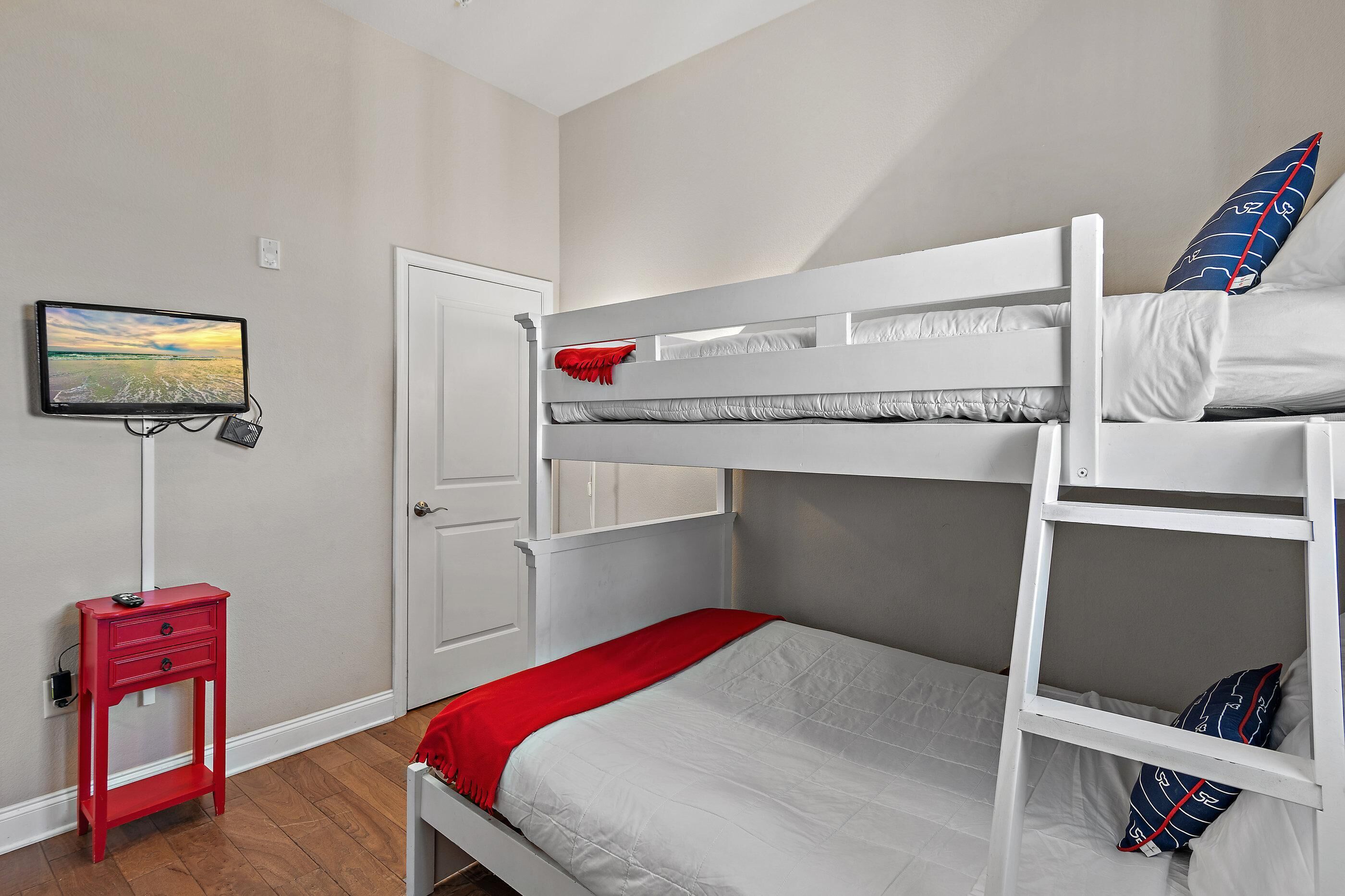 Un lit superposé avec un téléviseur fixé au mur, une table de chevet rouge et une porte visible au fond.