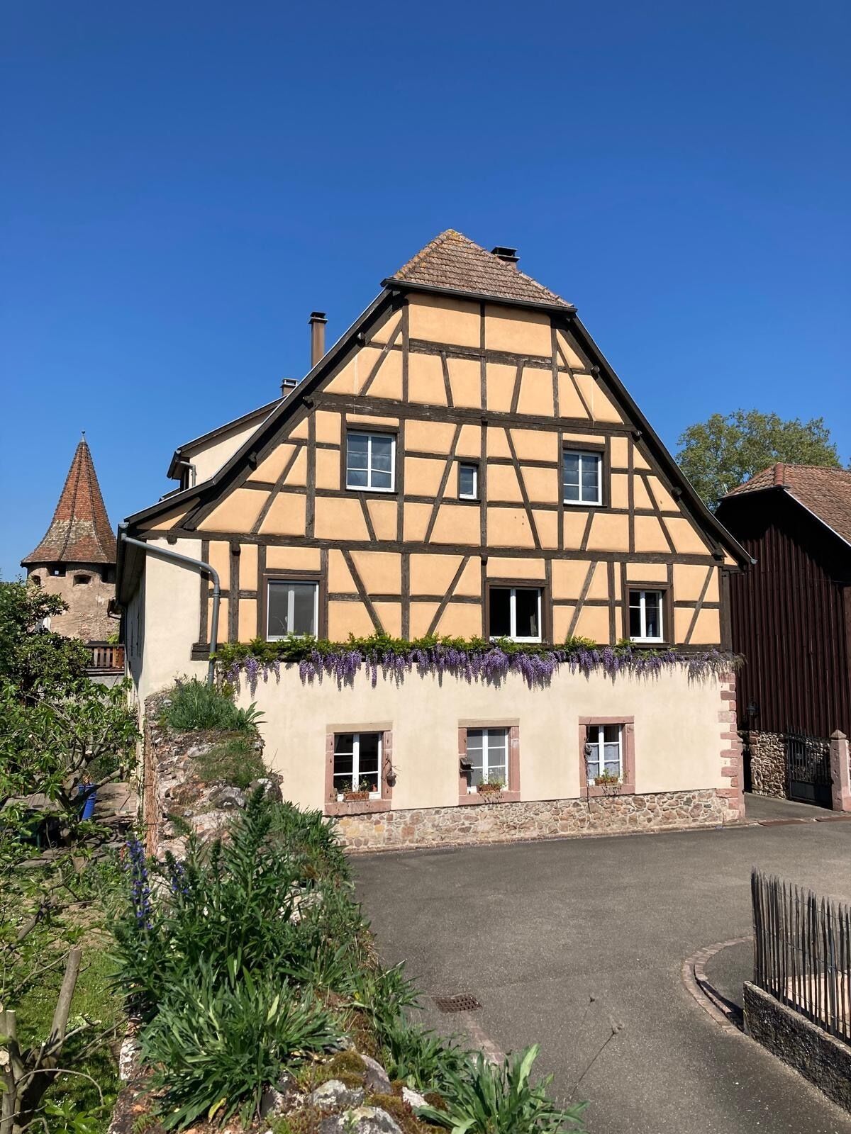 A traditional half-timbered house with a stone foundation, multiple windows, and a small garden area with purple flowers.