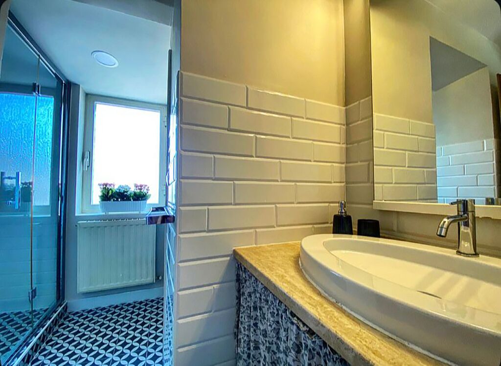Un baño con pared de ladrillo blanco, un lavamanos grande con espejo y una ventana con flores.