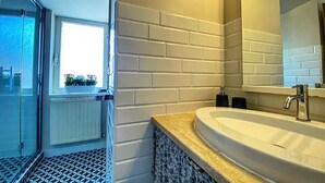 Ein Badezimmer mit weißer Ziegelwand, einem großen Waschbecken mit Spiegel und einem Fenster mit Blumen.
