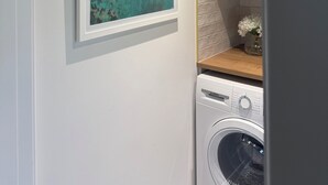 A laundry area with a washing machine, a shelf with baskets, and a framed painting on the wall. - Charming Newly Renovated 1 Bedroom Chalet | Stunning Sea Views (South Whiteness)