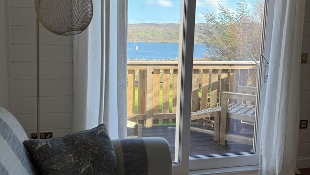 A room with a sofa, a hanging light fixture, and a sliding glass door leading to an outdoor deck with a wooden railing. - Charming Newly Renovated 1 Bedroom Chalet | Stunning Sea Views (South Whiteness)