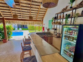 Zona de bar con vista a la piscina, un refrigerador lleno de bebidas y techo de paja.