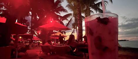 A beachside bar with outdoor seating, palm trees, and a view of the ocean.