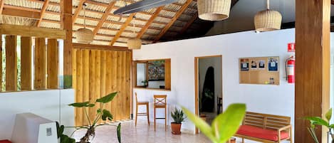 Una habitación con alto techo de madera, lámparas colgantes de mimbre, un banco de madera, una mesa y plantas en macetas.