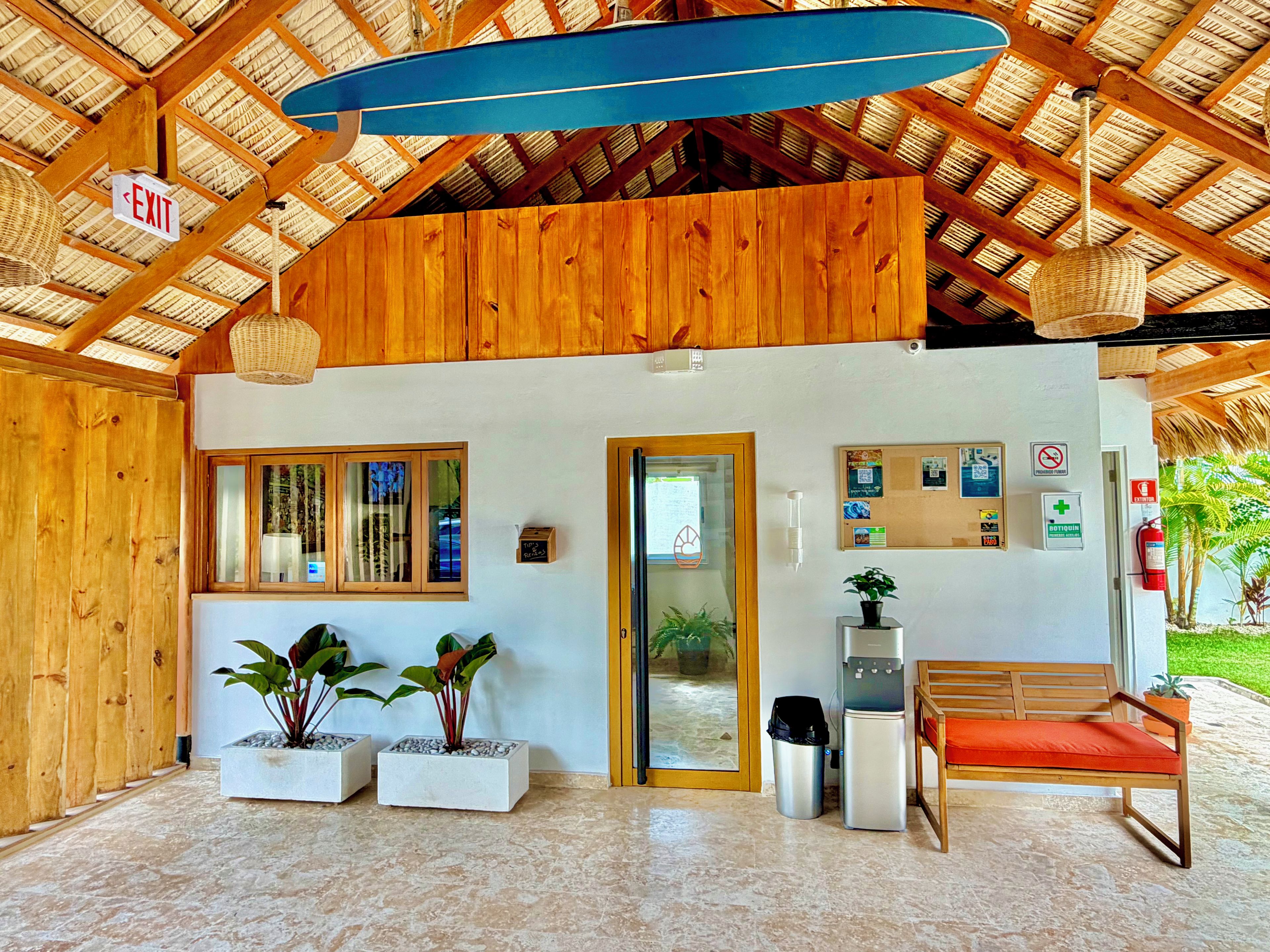 A reception area with a wooden ceiling, a reception desk, a bench, potted plants, a water dispenser, and a fire extinguisher.