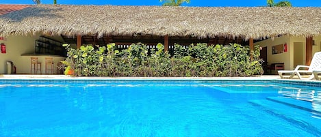 A swimming pool with a thatched-roof structure, a bar area, and a view of palm trees.