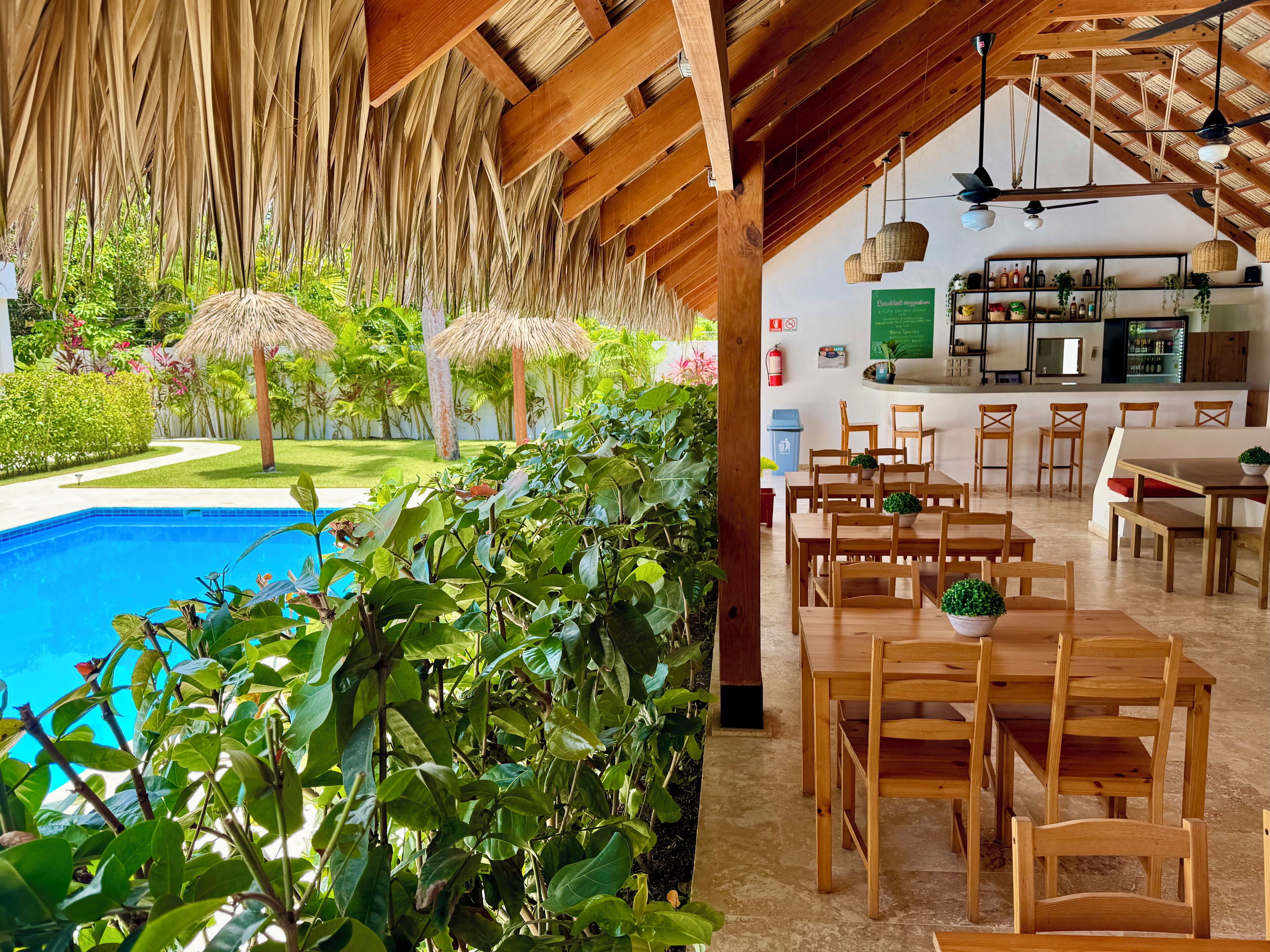 A poolside area with a wooden deck, outdoor seating, and a bar area with shelves of bottles and plants.