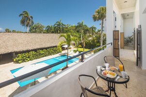 A balcony with a pool, dining table, and chairs, overlooking a pool area with a thatched roof and palm trees.
