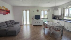 Een ruime woonkamer met een bank, een eettafel en een keuken met kastjes en een televisie.