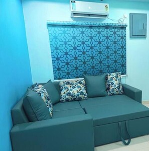 A room with a teal sofa, patterned cushions, and an air conditioning unit on the wall.