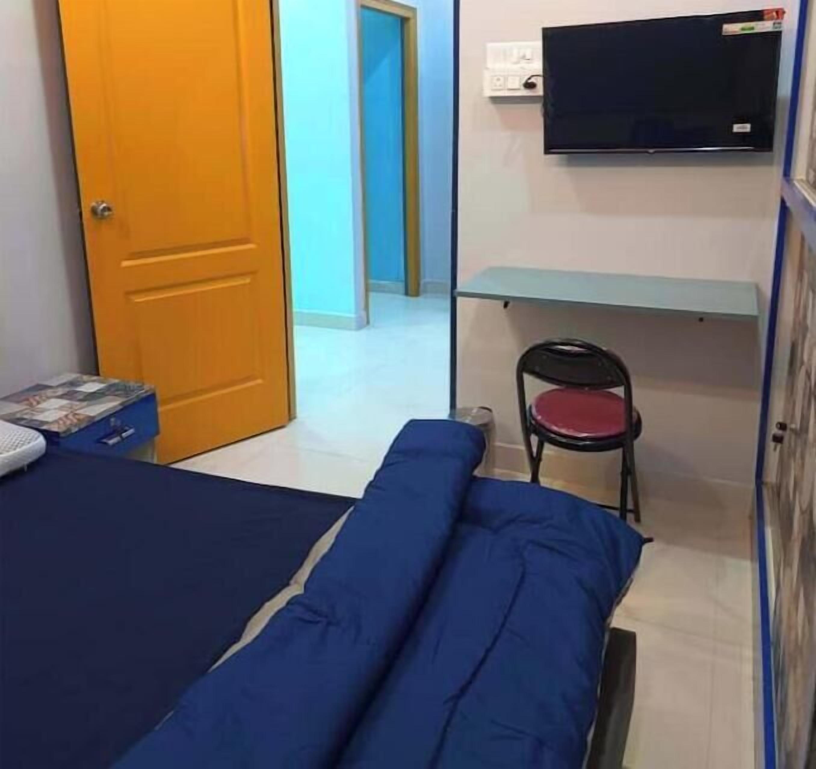 Een kleine kamer met een bed, een stoel, een televisie aan de muur en een deur naar een andere kamer.