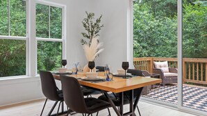 A dining area with a wooden table set for four, surrounded by black chairs. There is a large window on one side and a view of a deck with a checkered floor.