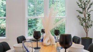 A dining table set with white plates, bowls, and black wine glasses, surrounded by chairs, with a large window and greenery visible outside.