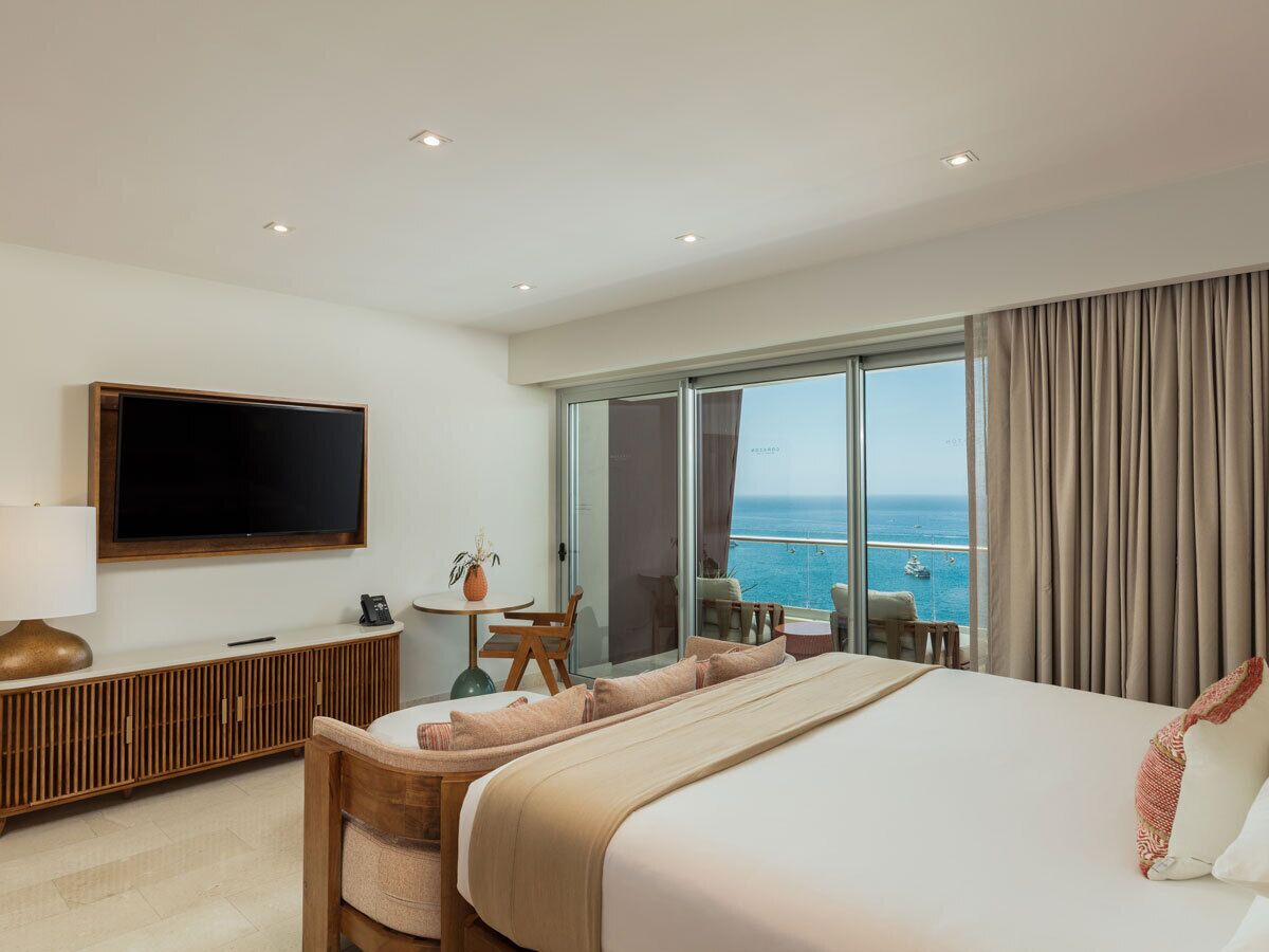 A hotel room with a large bed, a TV, a small table with chairs, and a balcony with a view of the sea.