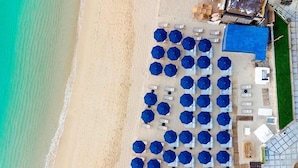 Une vue aérienne d’une plage où l’on aperçoit des rangées de parasols bleus et de chaises longues blanches, ce qui indique la présence d’un complexe hôtelier en bord de mer.