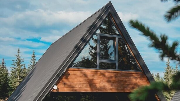 Une cabane moderne en forme de « A », dotée d’une grande fenêtre, d’une façade en bois et d’un espace extérieur aménagé pour s’asseoir.
