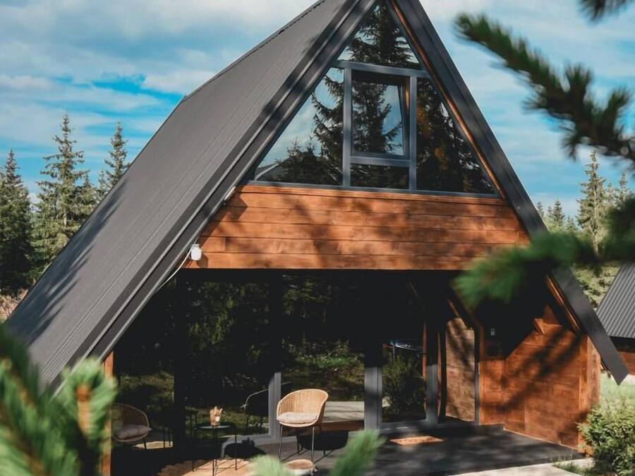 A modern A-frame cabin with a large window, wooden exterior, and outdoor seating area.