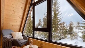 L’intérieur d’un chalet accueillant, avec une grande fenêtre, des murs en bois, un fauteuil à bascule, une petite table et une lanterne.