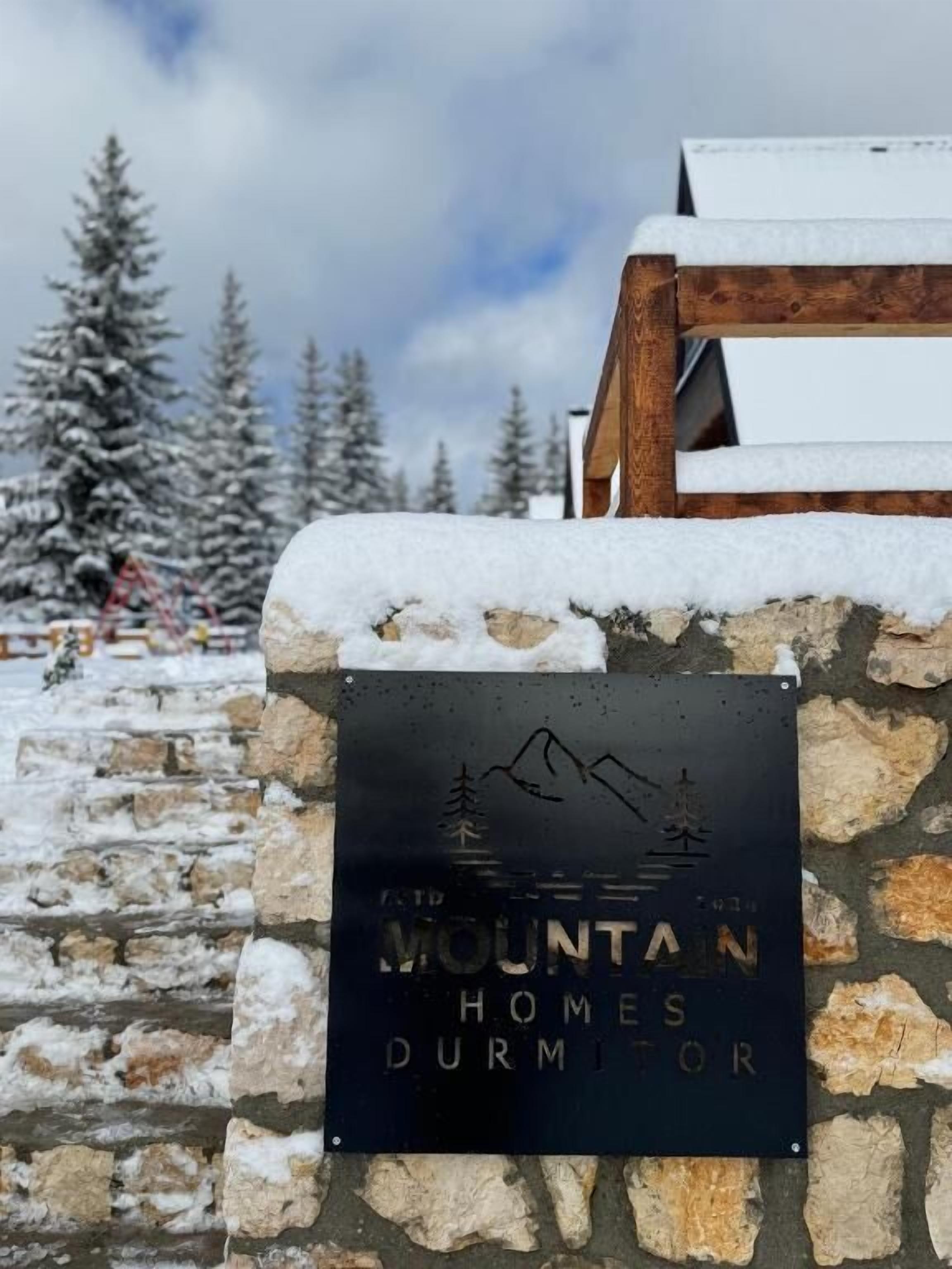 Letrero de 'Mountain Homes Durmitor' con un gráfico de una montaña y un árbol, sobre un muro de piedra con nieve.