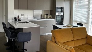 Eine moderne Küche mit grauen Schränken, einem Edelstahlherd und einem eingebauten Ofen. Ein Essbereich mit Tisch und Stühlen, sowie eine Couch im Vordergrund.