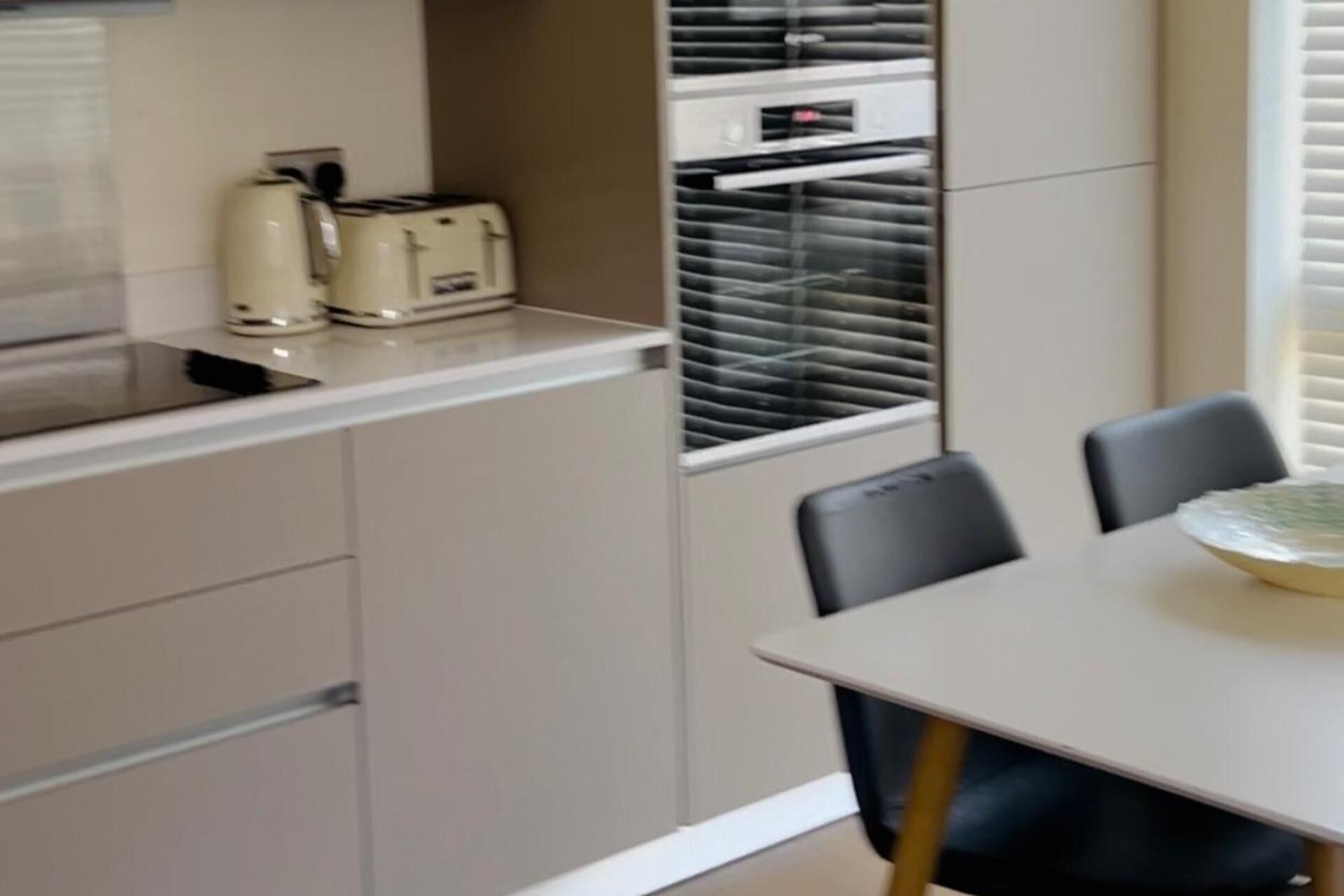 A kitchen with a built-in oven, a toaster, and a window with blinds. There is a table with chairs and a bowl on it.