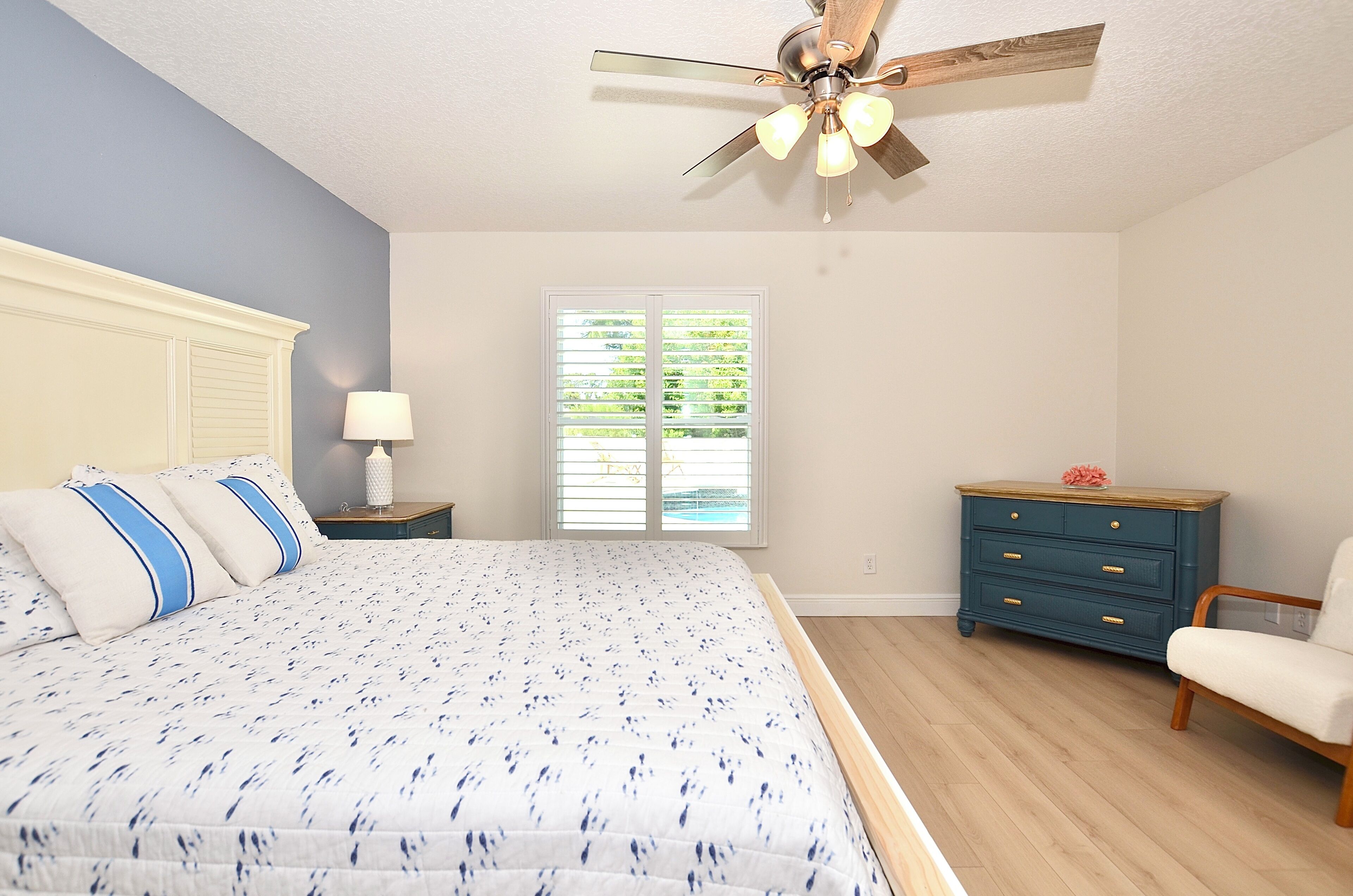 Um quarto com cama, mesa de cabeceira, cômoda, cadeira e ventilador de teto.