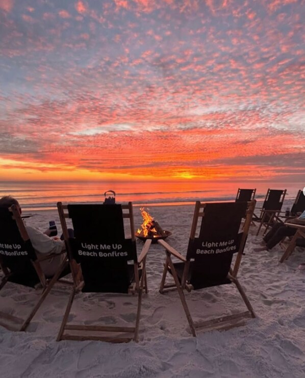 Beach chairs with 'Light Me Up Beach Bonfires' signs, a fire pit, and a sunset view.