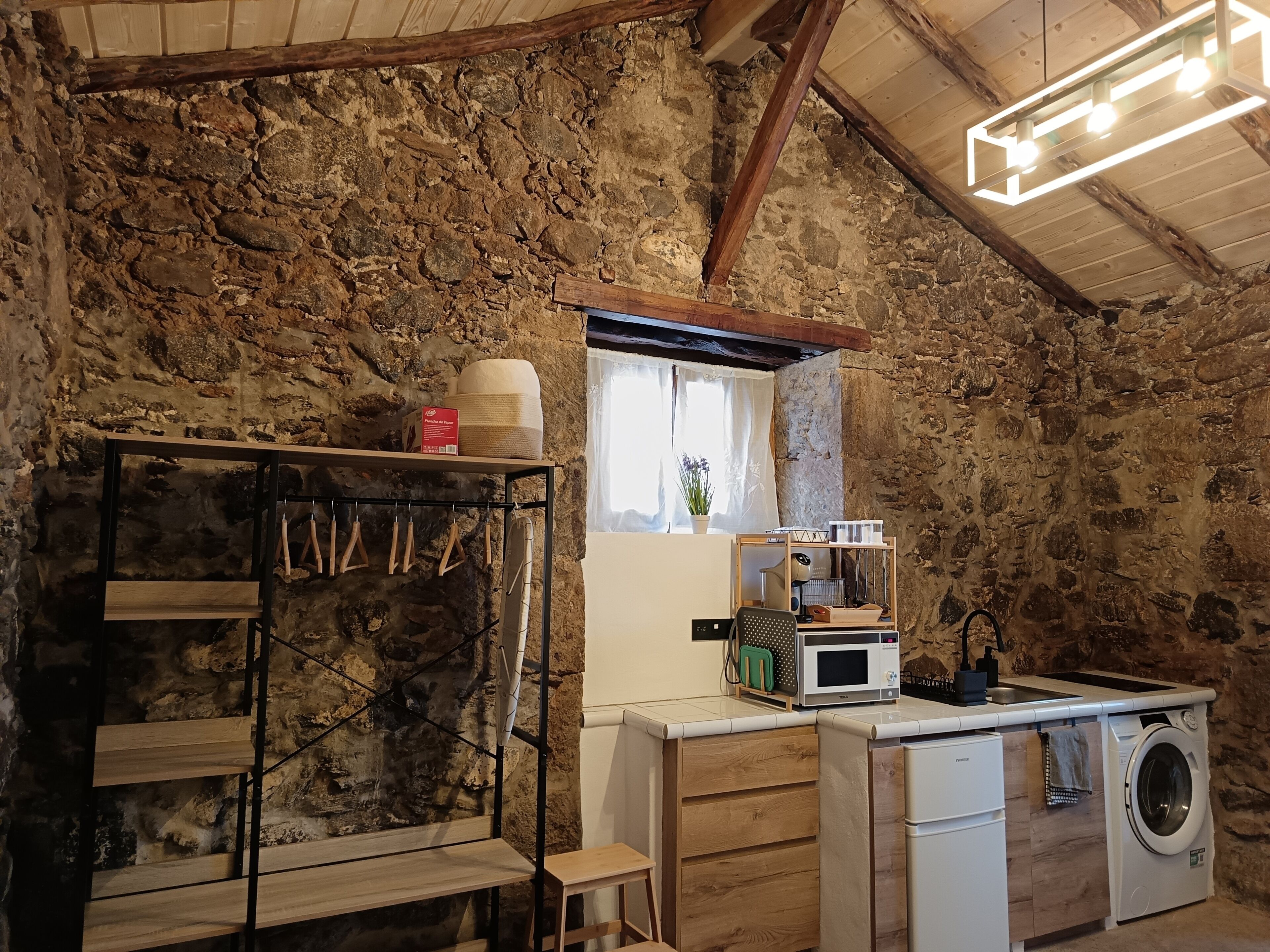 Uma cozinha rústica com paredes de pedra, vigas de madeira, máquina de lavar embutida e um balcão compacto com micro-ondas.